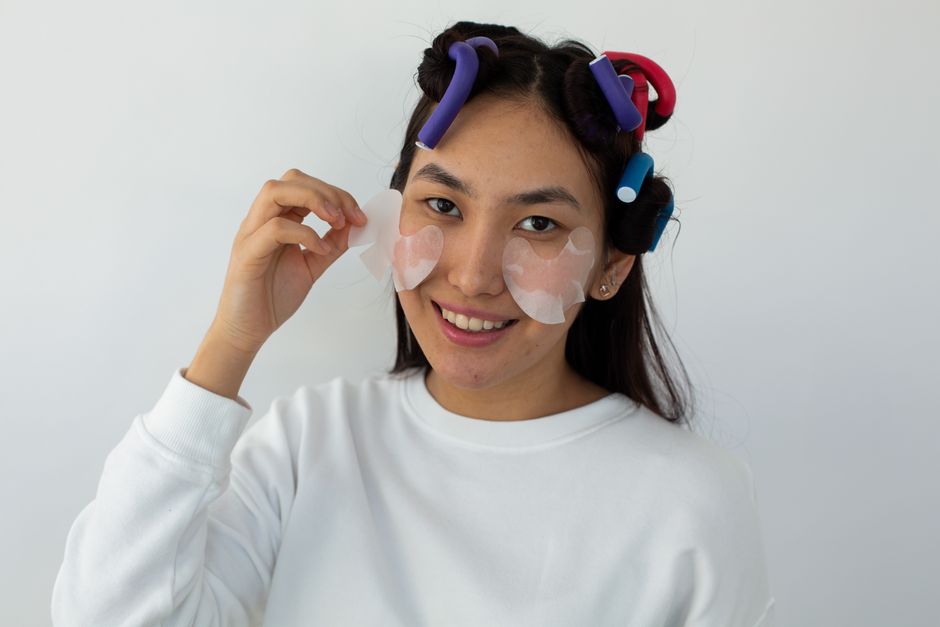 A woman applies under-eye masks with hair curlers during a skincare routine.