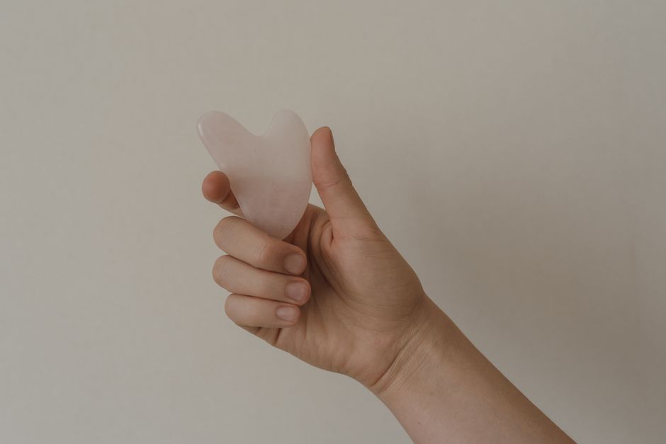 A hand holds a rose quartz gua sha tool against a neutral background, showcasing skincare elements.